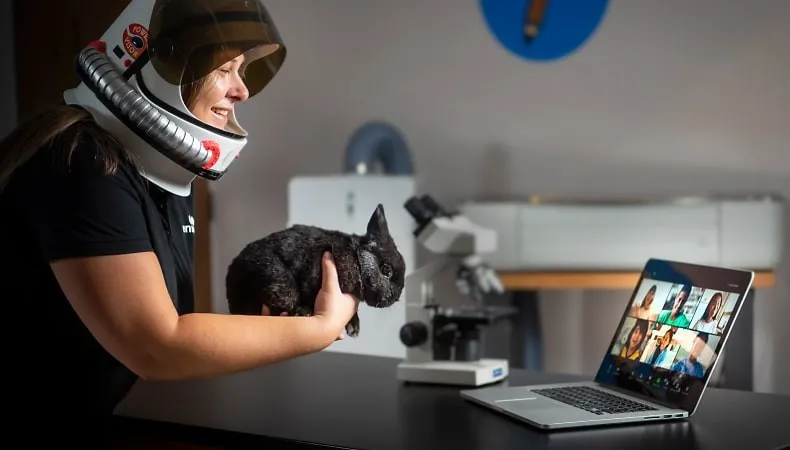 A woman wearing an astronaut’s helmet holds a rabbit up to the camera of a laptop, which is open in front of her. Children’s faces are visible on the laptop screen via videoconferencing.