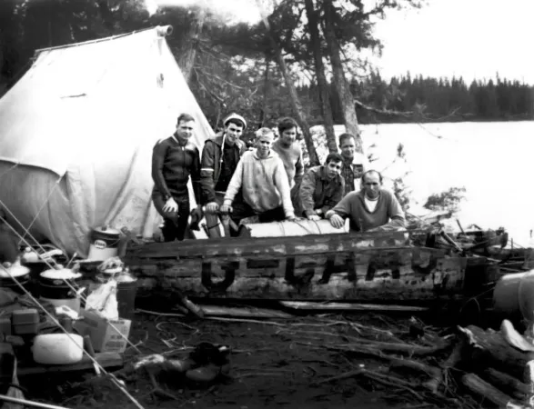Une image en noir et blanc présente un groupe de sept hommes debout derrière un gros morceau de bois sur lequel sont peintes les lettres d’appel « G-CAAC ». Ils se tiennent près d’un lac. Derrière eux se trouvent une tente blanche et un petit groupe de trois arbres. Devant eux, du côté gauche de la photo, on voit une pile d’équipement de camping et de cuisson. 