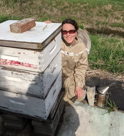 A woman wearing a protective jumpsuit crouches next to a beehive.