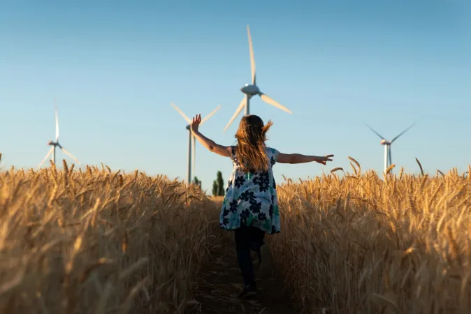 A young girl wearing a flowered dress is running through a path in a wheat field. Wind turbines are visible in the background, against a bright blue sky.