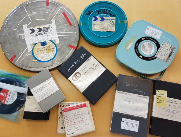 A variety of audio-visual formats, including film, audio, and different kinds of video cassettes, from the Les Harris Fonds are spread out on a table. 