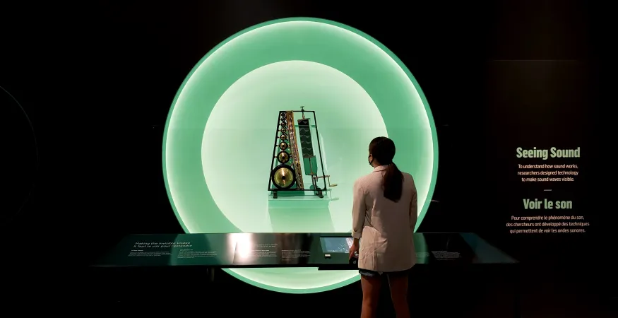 A woman is silhouetted in front of a circular, glowing showcase presenting the Koenig Sound Analyser. The title, “Seeing Sound” is visible on the wall.