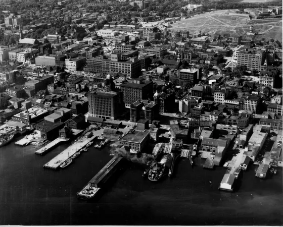 A black and white photograph taken from the air. The photo shows a series of docks with buildings on the port, and city streets behind. The Halifax Citadel is in the background.