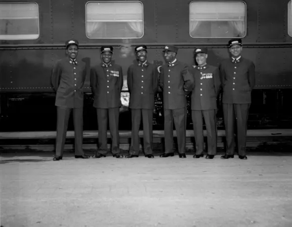 Black and white photograph of 6 uniformed Black porters standing in a line in front of a train. Some porters are smiling and looking at each other.