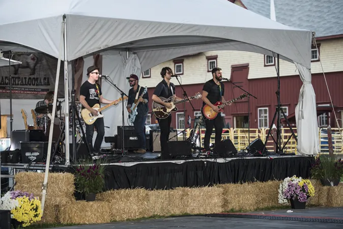 A tent set up on the side of a pavilion with five members of a band performing. Multiple microphones and amplifiers are set up on the stage and several haystacks are lined up along the front of the stage.