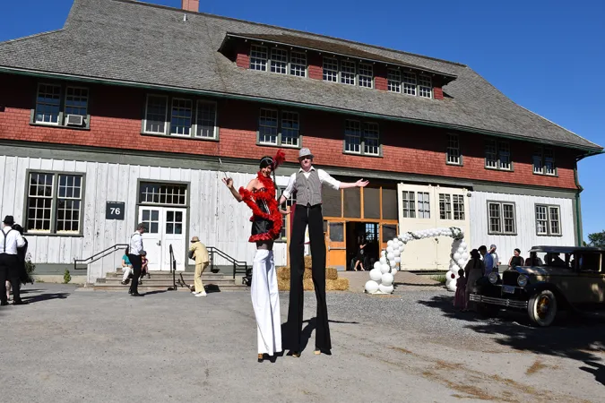Two people standing on stilts outside a large red a white barn. A balloon arch frames the door way of the barn and several people are pulling up in black cars and formal dresswear.