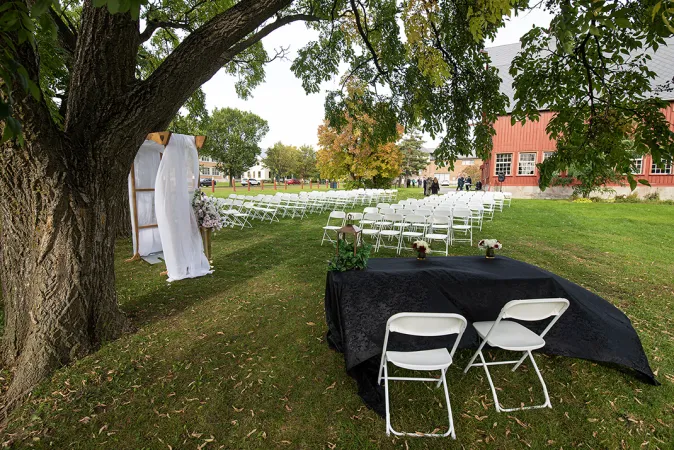 Lawn set up for a wedding with rows of foldout white chairs and an arch under a tree. A large red barn is visible in the background.