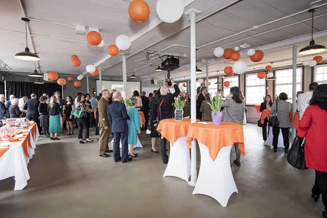 Groups of people standing around a large room with white ceiling and light walls and floors. There are small high tables scattered around the room decorated with orange tablecloths, flowers, and balloons