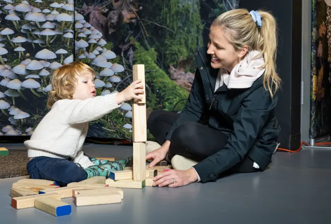 A young child plays with wooden blocks alongside a smiling adult with long blond hair tied back in a ponytail