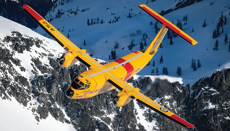 Yellow and red Buffalo CC-115 aircraft in flight, set against a snowy mountainous terrain.