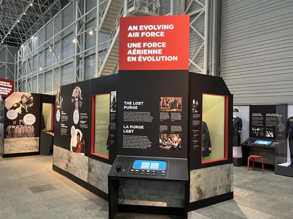 Several exhibition display cases, panels, and screens in a large, brightly-lit air hangar-style room with a high ceiling and tall, exposed metal walls.