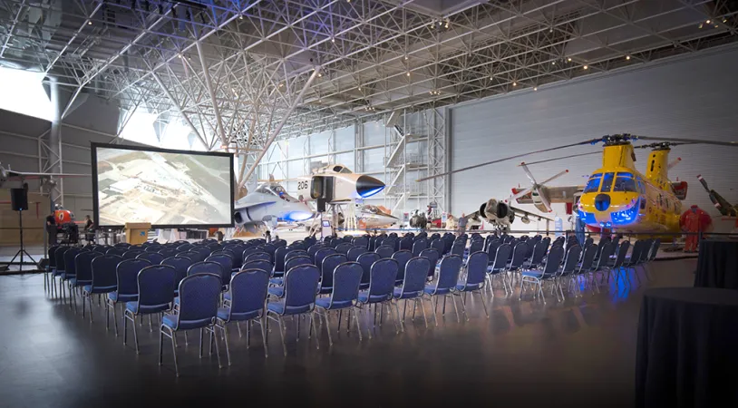 Des centaines de chaises sont alignées face à un grand écran, au centre d’une grande pièce. Un hélicoptère jaune est visible en arrière-plan, ainsi qu’une partie d’un petit avion blanc.