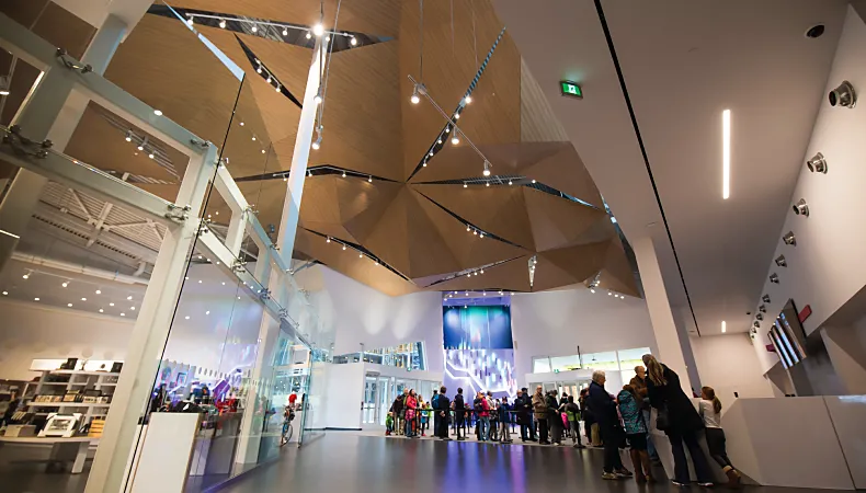 The front entrance of the Canada Science and Technology Museum. One side of the room features the welcome desks with screens above the desk. The ceiling is high light wooden geometric shapes with lights peaking through. The other side features a glass wall looking into the boutique.