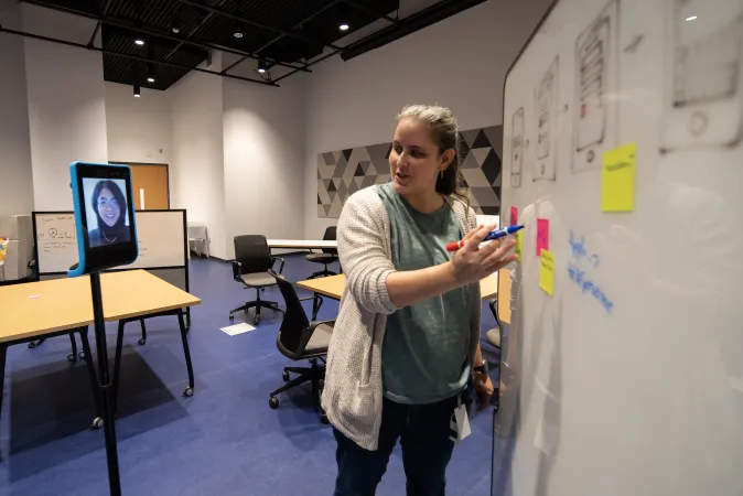 Two people talking by a whiteboard - one person is pointing towards a few sticky notes on the whiteboard. The other person is speaking through a screen on wheels that can be remotely controlled.