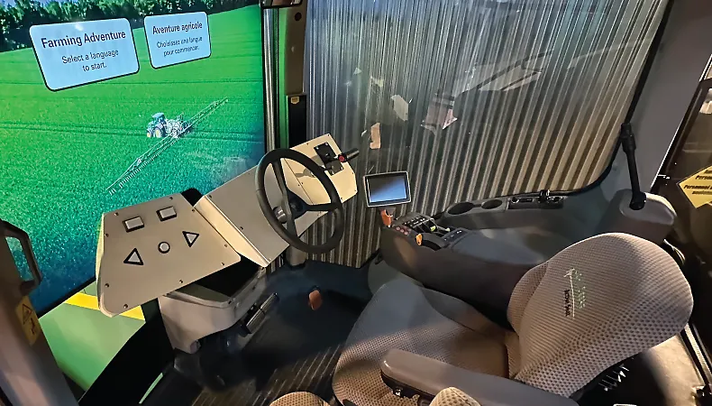 The interior of a museum interactive, which includes a chair, a steering wheel and a large screen showing a green field.