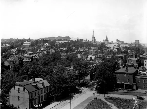 : A black and white photograph of a city street with houses and trees. There are church spires in the distance.