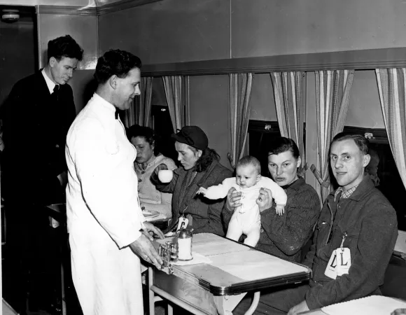 A black and white photograph of two men and two women sitting in a train car. One man holds an infant. Two men stand in front of them serving food or drink.