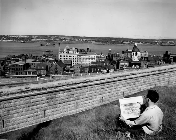 Une photo en noir et blanc d’un homme avec une tablette à croquis, assis sur une colline, regardant la ville. On voit le havre d’Halifax en arrière-plan.
