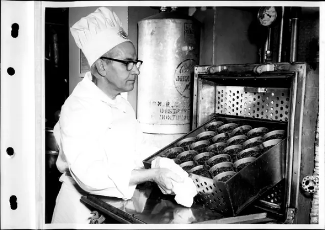 Image is a black-and white-photograph showing a man in a white chef’s uniform, holding a metal tray with puddings in metal molds.