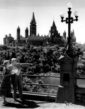A black and white photograph of a man and a woman pointing toward  a waterway, with the large, stone Parliament Buildings visible in the background.