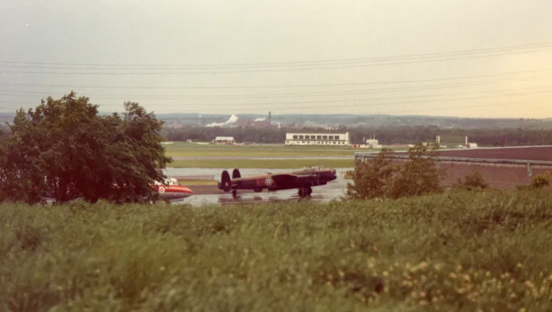 Looking down from a grassy area towards a building and aircraft. The Gatineau Hills are in the distance.