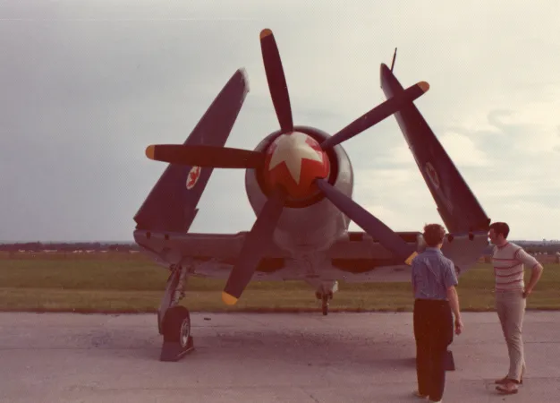 Front view of an airplane’s nose. The airplane’s wings are folded upward. Two men talk in the foreground.