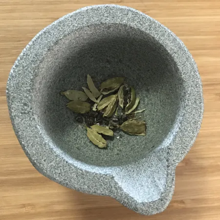 Un petit mortier de pierre grise contient six gousses de cardamome écrasées. Les graines libérées de leur enveloppe écrasée sont visibles.
