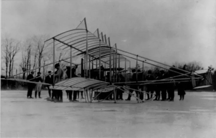 L’image est une photo noir et blanc montrant un biplan Herring Burgess sur la surface gelée d’un lac. Une foule est assemblée derrière l’avion. 