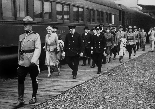 A black and white photograph of uniformed RCMP officer marching beside a train car, in front of an elderly man and woman, followed by several people in formal attire, followed by additional RCMP officers. 