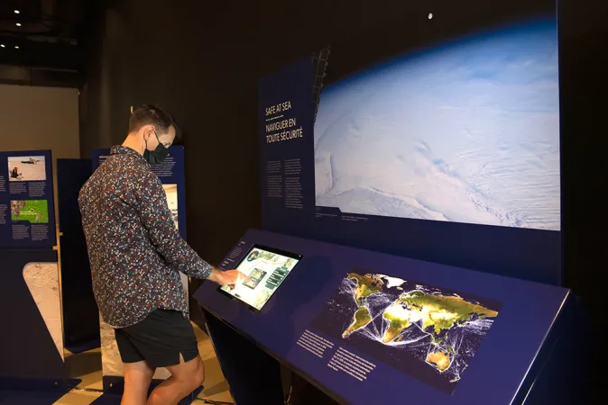 A person in a multi-coloured shirt and black shorts interacts with a touch screen on an exhibition module.  To the right of the screen, on the module, is a high contrast map of the world.  Above the module is a large white image.