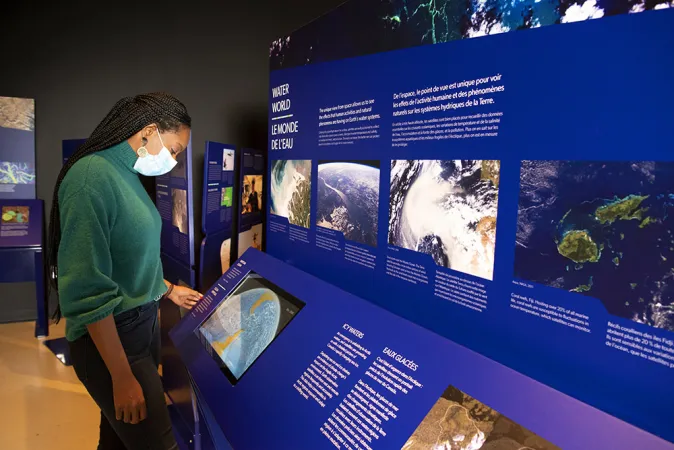 Une personne portant un chandail vert et un pantalon noir se tient devant un module d’exposition. Le module est bleu avec du texte blanc et comprend une série de photographies colorées de la Terre prises de l’espace.