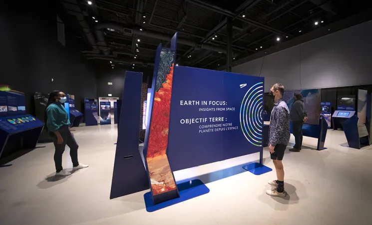 A few people are in a gallery with blue exhibition modules. One person is standing in front of a panel where the title of the exhibition; Earth in Focus: Insights from Space, can be read in large white print. To the right of the title is a graphic representation of the Earth, composed of concentric rings of blue, green and white. In white silhouette, a graphic image of a satellite can be seen in orbit.