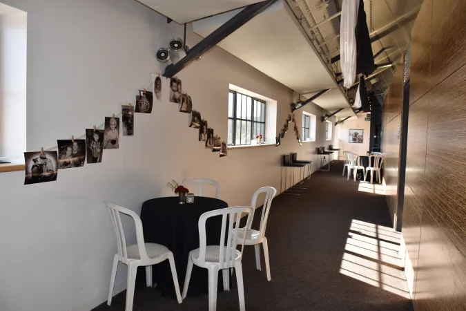 A long hallway with white and light wood tones. Several small tables with a couple chairs each are set up around the room. One wall is white and has several strings of photos hanging across.