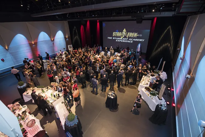 Top down view of a room decorated with various space-themed items and small circular black tables scattered around the room. Multiple tables are set up on one side of the room with food and drinks on the other side. On the projector screen, the words “Star Trek the startleet academy experience” is displayed. Many people in formalwear are standing in groups around the room.