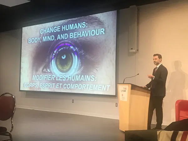 Eric Ward is dressed in a black suit and maroon tie. He stands behind a podium and microphone. Behind him is a projector screen with an image of an eye with a lens overlaid overtop. On the screen text reads “change humans: body, mind, and behavior”