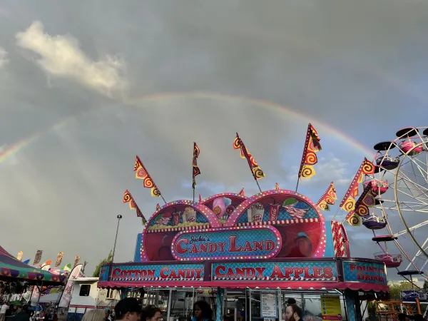 Un double arc-en-ciel encadre un kiosque de carnaval rose avec une grande roue en arrière-plan. 