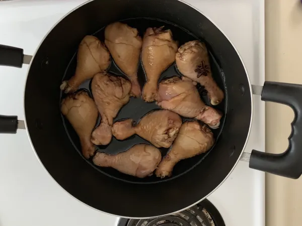 Des pilons de poulet cru sont disposés dans une casserole noire sur une cuisinière blanche. Les pilons sont brun pâle. Une fleur d’anis est visible sur l’un des pilons en haut à droite, et la casserole contient une petite quantité de liquide foncé.