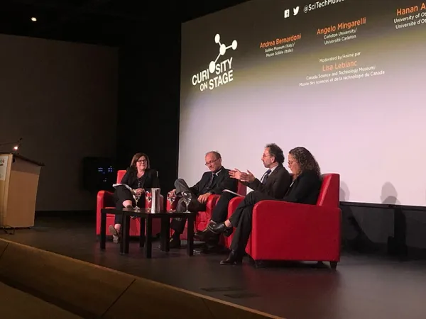 The four panelists are seated on a stage in red armchairs: Lisa Leblanc (moderator), Andrea Bernadoni, Angelo Mingarelli, Hanan Anis.  Angelo speaks and gestures with both hands as the others listen.