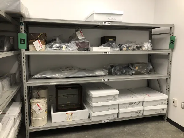 Grey metal shelving unit marked “B” which has radioactive artifacts stored on it. Artifacts are stored in white boxes on lower shelves, and are enclosed in clear plastic sheeting on the two upper shelves.