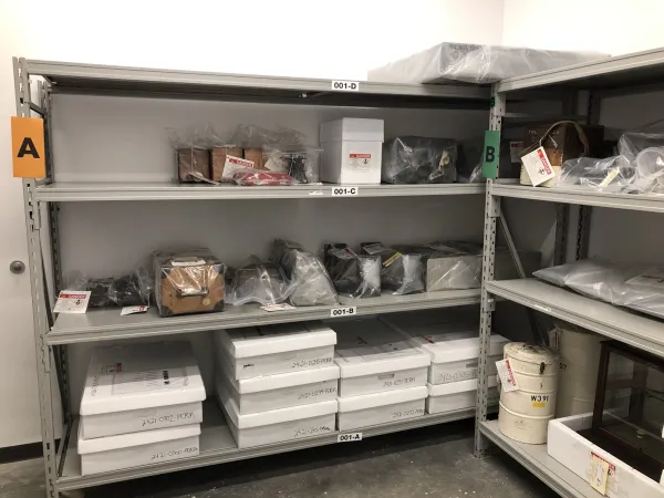 Grey metal shelving unit marked “A” which has radioactive artifacts stored on it. Artifacts are stored in white boxes on lower shelves, and are enclosed in clear plastic sheeting on the two upper shelves.