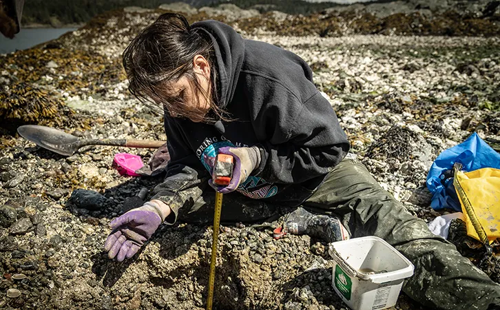 Photograph of a woman sitting on a rocky shoreline, using a measuring tape to measure the depth of a hole into the ground. She is wearing purple gloves, a black hoodie-style sweater, and pants.