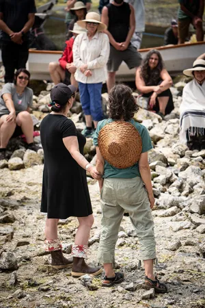 Photograph of two women, from behind. A women in a black dress holds the arm of another woman wearing a green shirt and beige pants, with a woven hat hanging down her back.