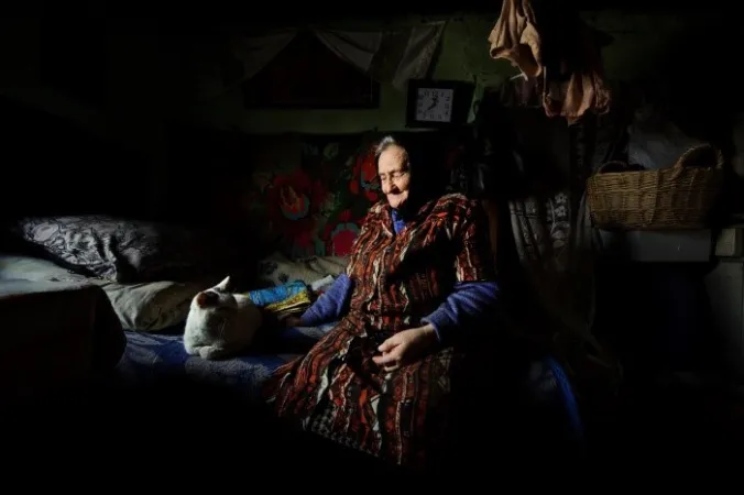 An old lady sits on the edge of a bed inside a dark room. A white cat is sitting next to her. There is a wicker basket behind her. A colourful tapestry hangs on a wall beneath a clock. 
