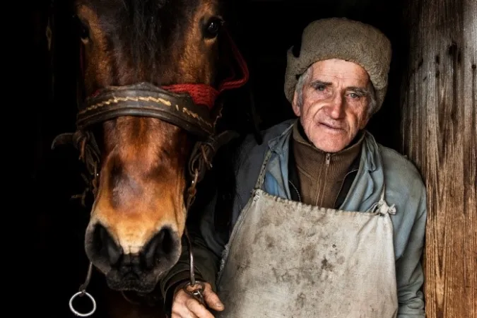 A close up portrait shot of an old farmer and his horse standing beside a wooden doorway. 
