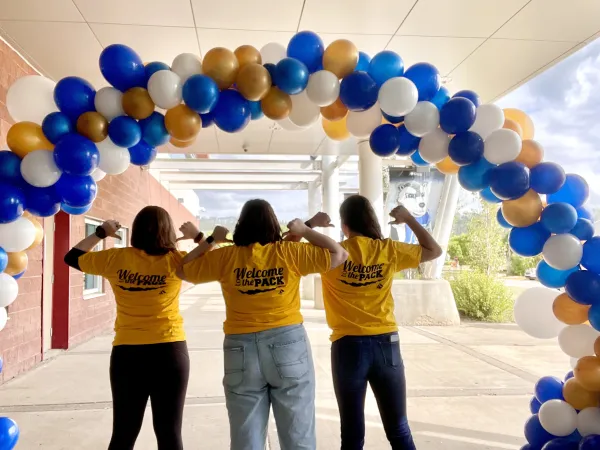 Three people in yellow shirts stand under an arch made of blue, gold, and white balloons. 