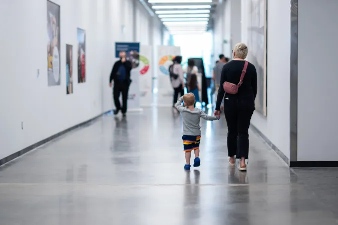 Distant view of people walking at the Ingenium Centre: a woman holding a child's hand seen from behind, a man approaching from the opposite direction, and posters in the background.