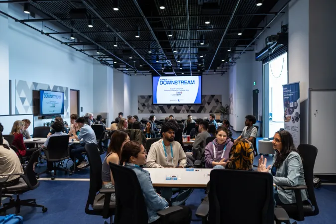 Une grande salle avec les mots « Downstream Networking » projetés par un projecteur le long d'un mur. De nombreuses personnes sont assises autour de diverses tables et discutent.