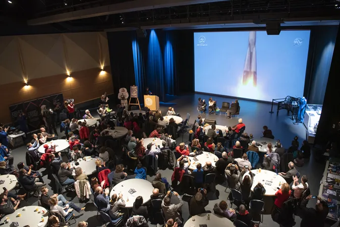 Multiple big white circular tables are set up around a large room with several people sitting at each one. At the front of the room, three people are sitting in large chairs speaking and pointing at a screen where an image of a rocket launch can be seen.