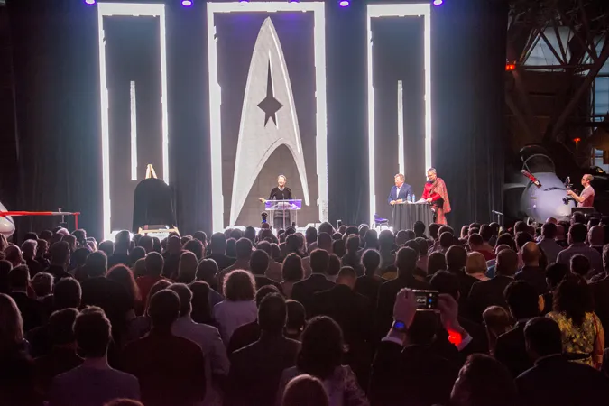 A large crowd of people facing a stage with black curtains. Three people are standing on the stage speaking to the audience and behind them are bright silver lights in different patterns, one of which is the Star Trek symbol. 