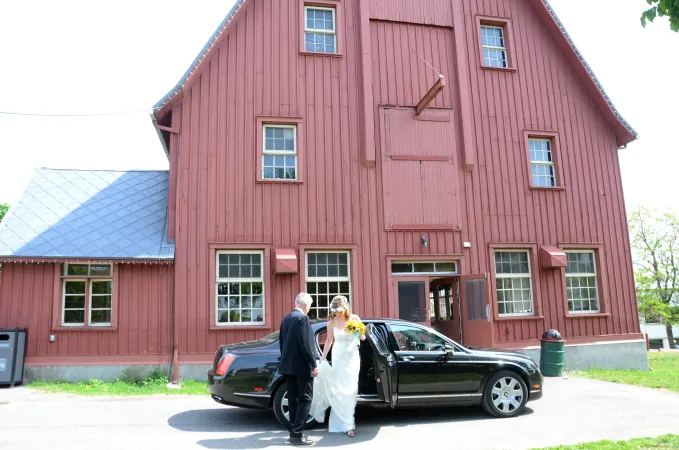 A bride getting out of a black car in front of a big red barn. A second person is holding the train of the white dress.